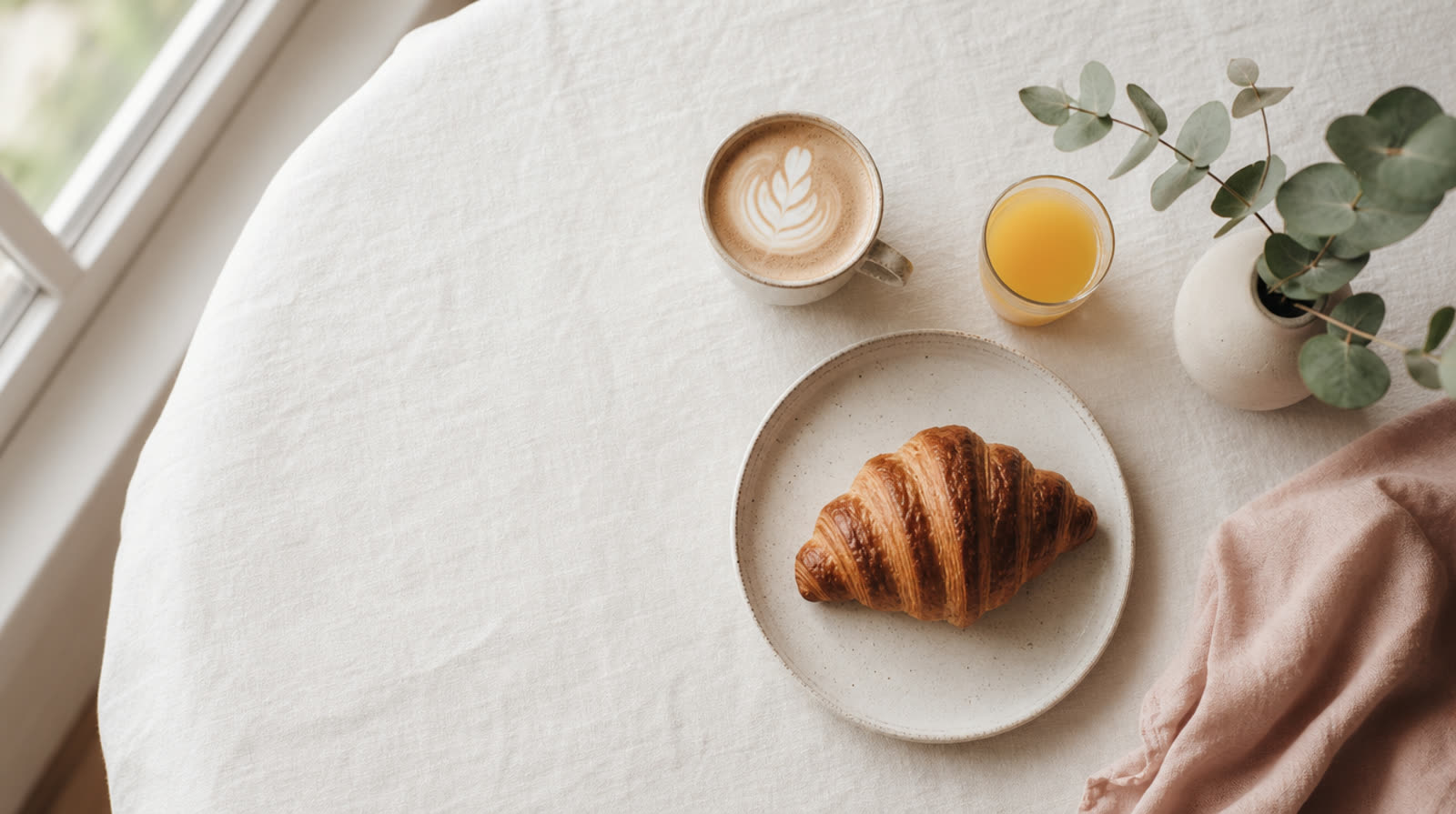 A brunch table from above — a latte, a glass of orange juice, a croissant and a sprig of eucalyptus on linen